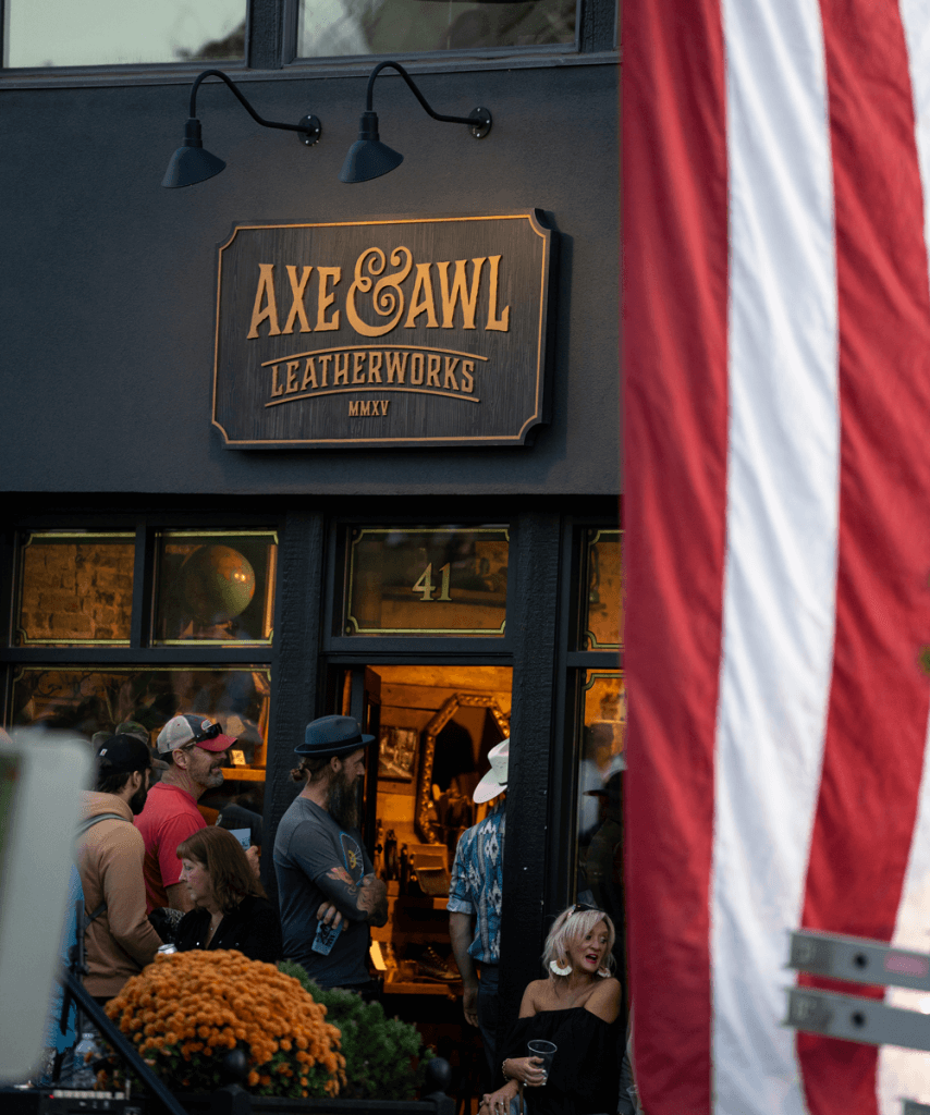 The brick and mortar leather shop of axe and awl leatherworks with waynesville locals sitting out front under the american flag. 
