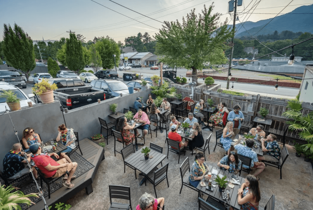 Out back of Axe and Awl Leatherworks as it operates as Bevel Bar, full of people enjoying a beautiful evening with the Smoky Mountains as the backdrop