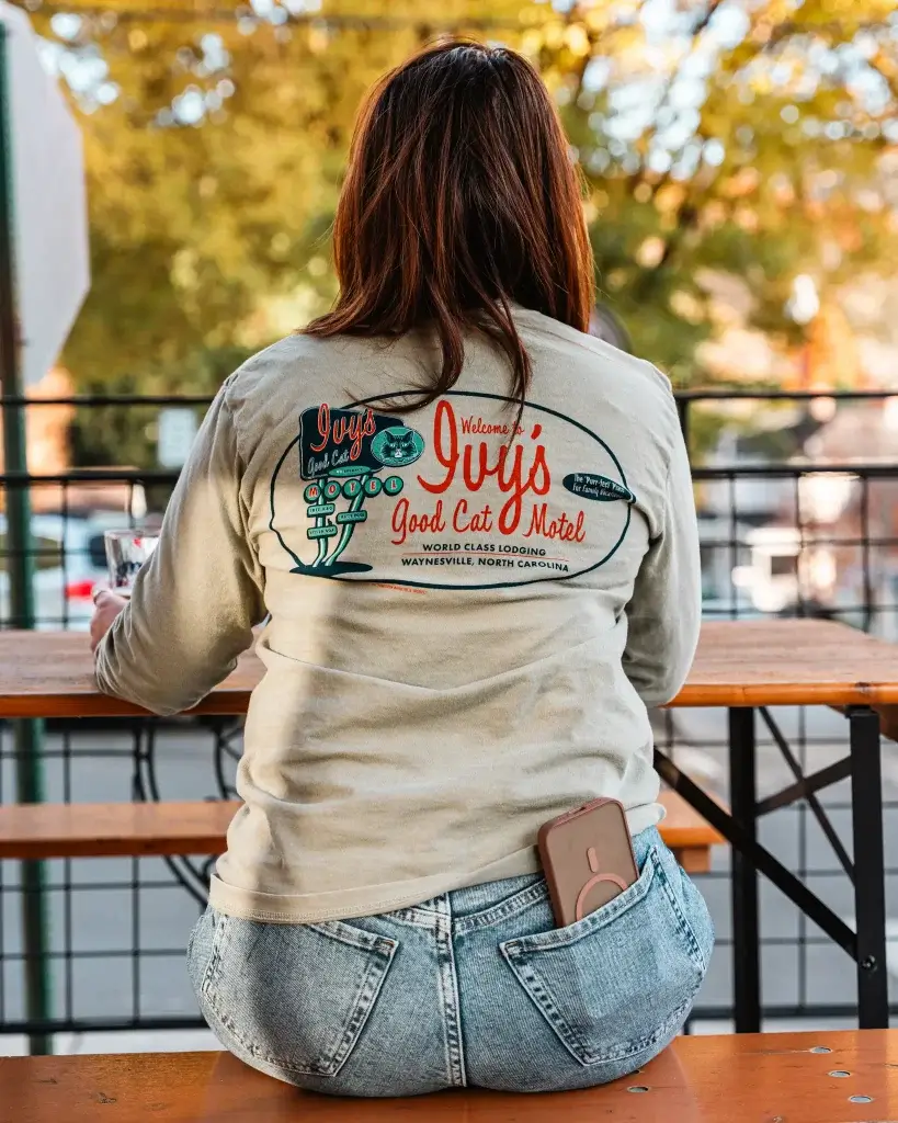 a red haired woman sits at a picnic table in downtown waynesville, nc wearing a long sleeve ivy's good cat motel ringer tee